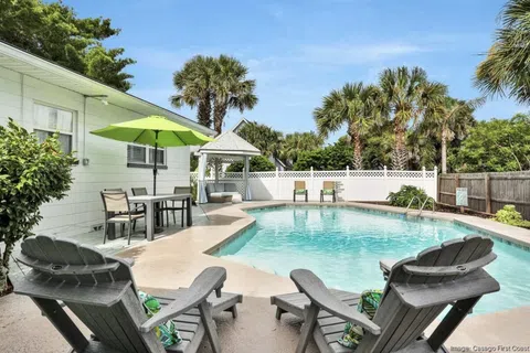 Sunny backyard pool with lounge chairs, green umbrella, gazebo, and palm trees under a blue sky.