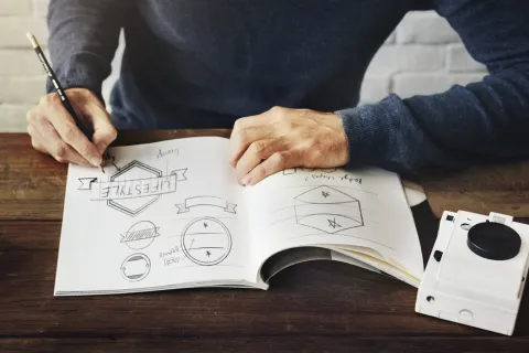 Designer sketching logo concepts on a notebook at a wooden desk with a vintage camera nearby