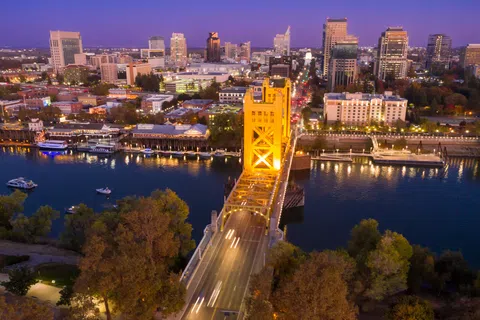 Aerial view of Sacramento city skyline at dusk with Tower Bridge lit and cars crossing over the river.
