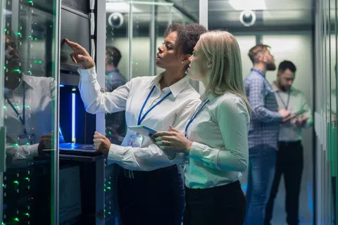 Two female IT professionals working and monitoring servers in a data center with two male colleagues in the background.
