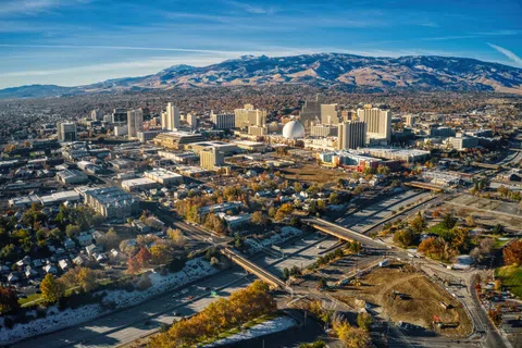 Aerial view of a mid-sized city with mountains in the background under a clear blue sky, showing urban buildings and roads.