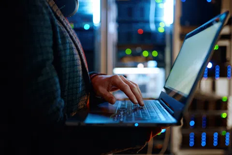 Person typing on laptop in server room with glowing network equipment in the background at night