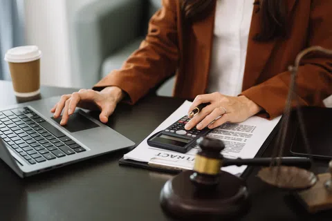 Person working on laptop and calculator with legal contract, gavel, and scales on desk symbolizing law and finance