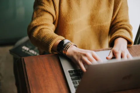 Person in mustard sweater working on a laptop with bracelets on wrist at a wooden desk.