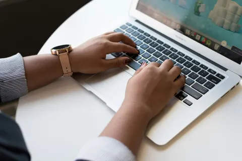 Person wearing watch typing on a silver laptop keyboard on a white desk in natural light.