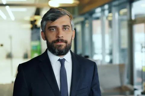 Confident businessman with gray beard wearing a dark suit and tie in a modern office setting.