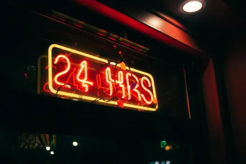 Red and yellow neon sign displaying 24 HRS inside a dimly lit building window with decorative lights.