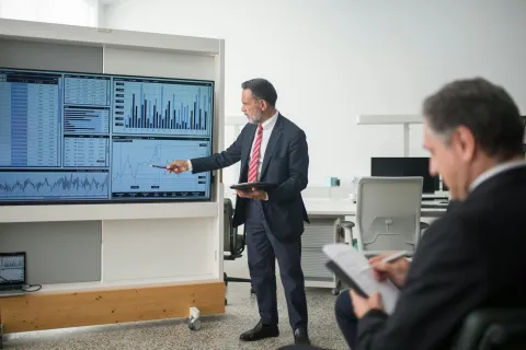 Businessman presenting financial charts and graphs on large screen while colleague takes notes in office.