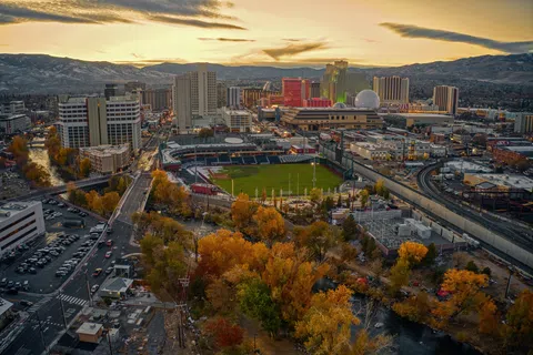Aerial view of a cityscape with a baseball stadium, autumn trees, and buildings under a cloudy sunset sky.