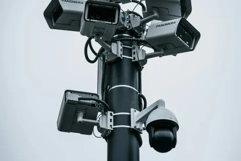 Multiple security cameras mounted on a tall black pole under a cloudy sky for surveillance.