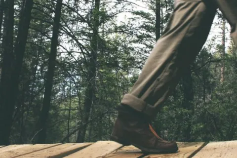 Close-up of a person walking on wooden path in forest wearing brown boots and rolled-up pants.