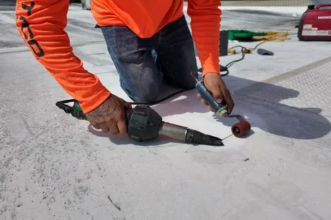 Worker in orange shirt kneeling on concrete using tools for floor repair or sealing outdoors under sunlight