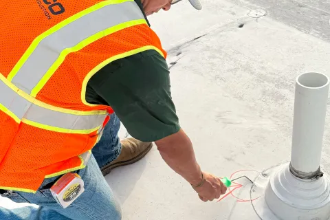 Construction worker in orange safety vest marking a spot on a rooftop near a white pipe vent.