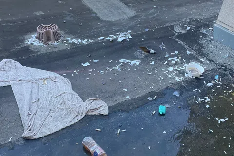 Broken glass, empty bottles, cigarette butts, and a towel scattered on a wet rooftop with greenery in background