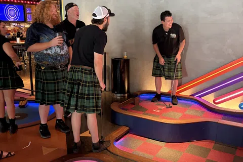 Group of people wearing kilts playing indoor mini golf under colorful neon lights in a casual setting.
