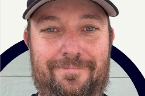 Smiling man with beard wearing a black Men In Kilts cap and shirt with a circular design background.