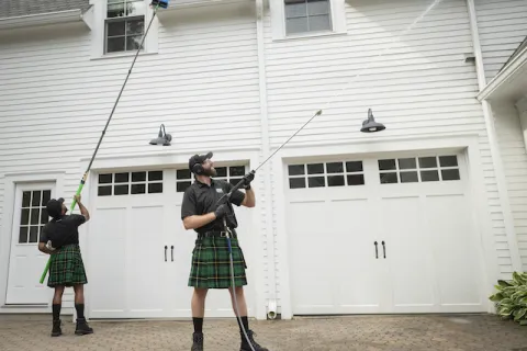 Two workers wearing kilts power washing exterior of white house with garage doors on driveway