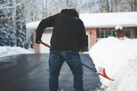Person shoveling snow from a driveway in front of a house on a clear winter day.
