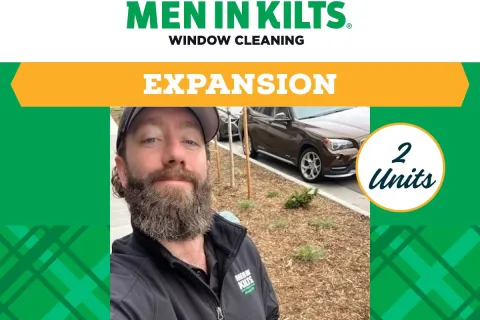 Man with beard wearing Men in Kilts jacket posing outdoors near parked cars and plants in Denver.
