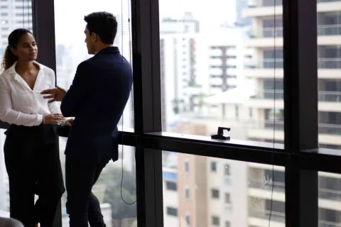 Two business professionals having a discussion by large office windows with urban cityscape in the background.
