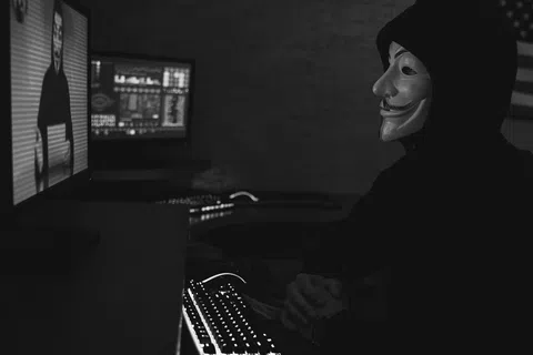 Person wearing Guy Fawkes mask and hoodie working on computer in dark room with multiple screens.