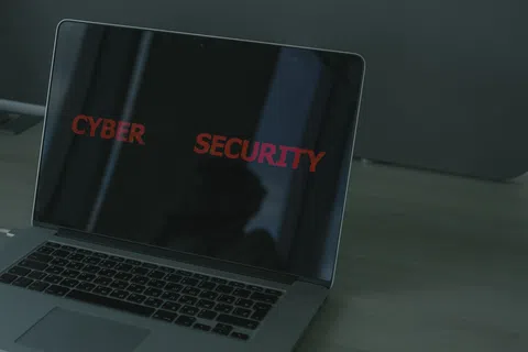 Laptop screen displaying red text Cyber Security on a dark background on a wooden desk.
