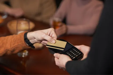 Person making contactless payment with a credit card and card reader in a casual setting.