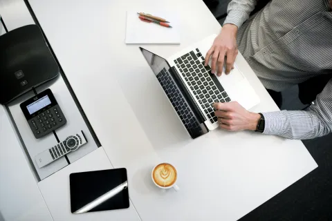 Hands typing on a laptop on a white desk with coffee, tablet, phone, notepad, and pens in office setting