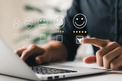 Person selecting five-star happy feedback on a digital rating scale with smiley faces above a laptop keyboard.