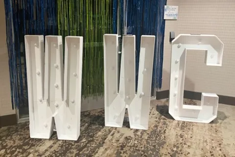 Large white marquee letters W, W, and C with bulbs standing on carpeted floor against colorful backdrop.