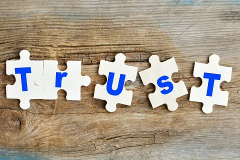 Wooden puzzle pieces spelling the word TRUST with blue letters on a rustic wooden background.