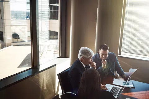 Three businesspeople in a meeting discussing documents and laptop in a sunlit office room near windows.