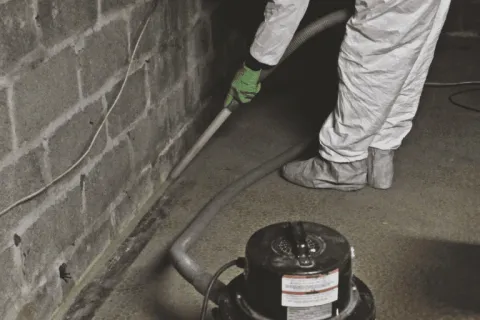 Person in protective suit using industrial vacuum to clean basement corner with concrete block walls.
