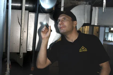 Technician in Mold Medics uniform inspecting ventilation ducts with flashlight in a dim basement.