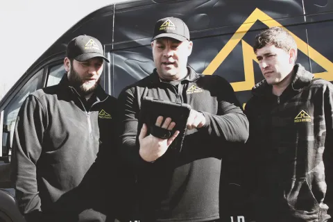 Three workers in black Milo Homes uniforms review a tablet in front of a branded service van under daylight.