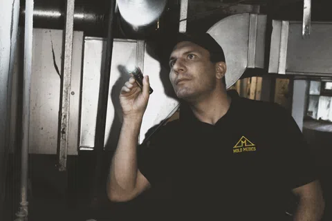 Technician inspecting HVAC system with flashlight in a basement wearing Mold Medics uniform and cap.