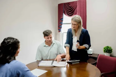 Three people in a meeting discussing documents at a round wooden table in a bright office.