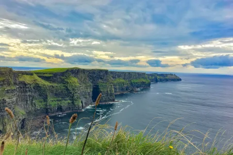 Scenic cliffs covered in green grass overlooking a calm ocean under a cloudy sky at sunset.