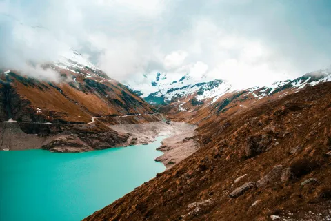 Turquoise mountain lake surrounded by rocky slopes and snow-capped peaks under a cloudy sky.