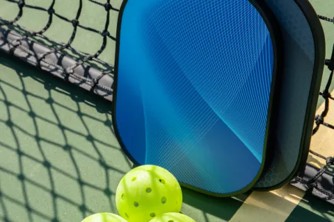 Three yellow pickleball balls and two blue paddles leaning against a net on a green pickleball court.