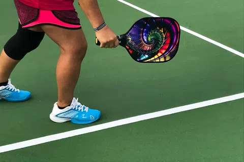 Player wearing blue shoes and pink skirt holding colorful pickleball paddle on green court near white lines