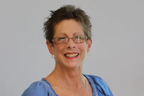 Smiling middle-aged woman with short hair and glasses wearing a blue blouse and gold necklace.