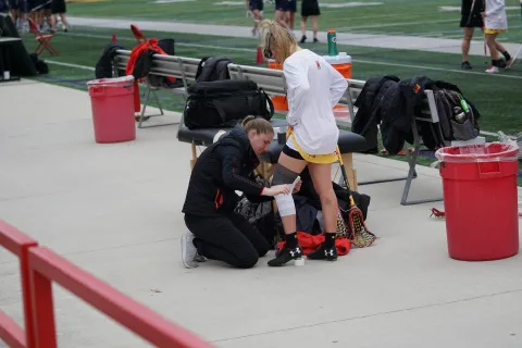 Athletic trainer wrapping tape on female lacrosse player’s knee near sports bench on field sideline.