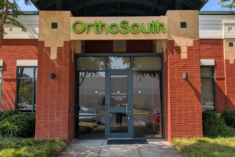 Entrance of OrthoSouth building with glass door, brick walls, greenery, and a curved black awning under a clear blue sky.