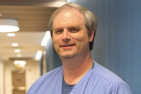 Male healthcare worker in blue scrubs writing on a clipboard in a hospital hallway with a phone in pocket.