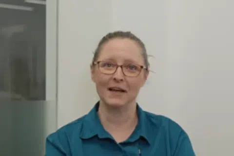 Woman with glasses and teal shirt speaking inside a modern office with glass walls and white background.