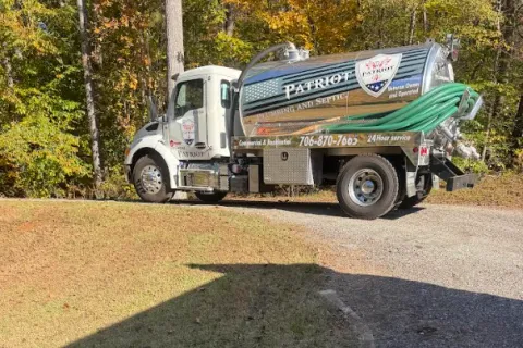 Patriot Plumbing and Septic service truck with hoses parked on a gravel driveway surrounded by trees in autumn.