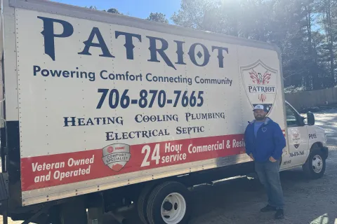 Man standing next to Patriot service truck advertising heating, cooling, plumbing, electrical, septic services and 24-hour support.