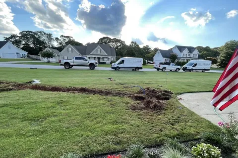 Residential lawn with trench dug for installation, parked service vans and trucks in background under partly cloudy sky.