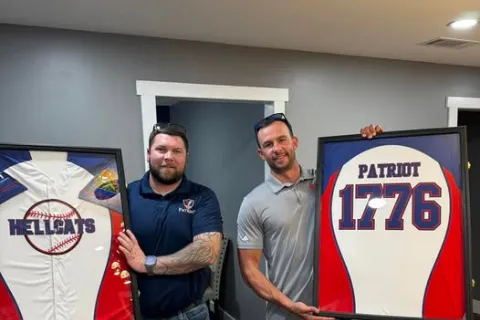 Two men holding framed baseball jerseys, one with Hellcats logo, the other with Patriot 1776 design inside a room.